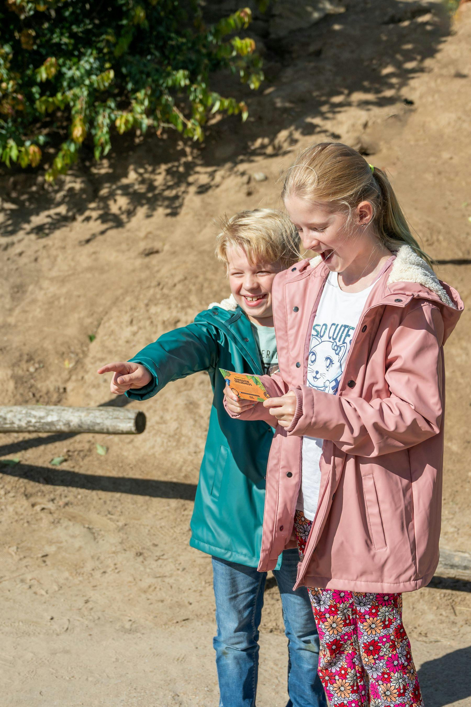 Two children smile and laugh outdoors. One child points ahead while the other holds a map. A dirt path and greenery are in the background.