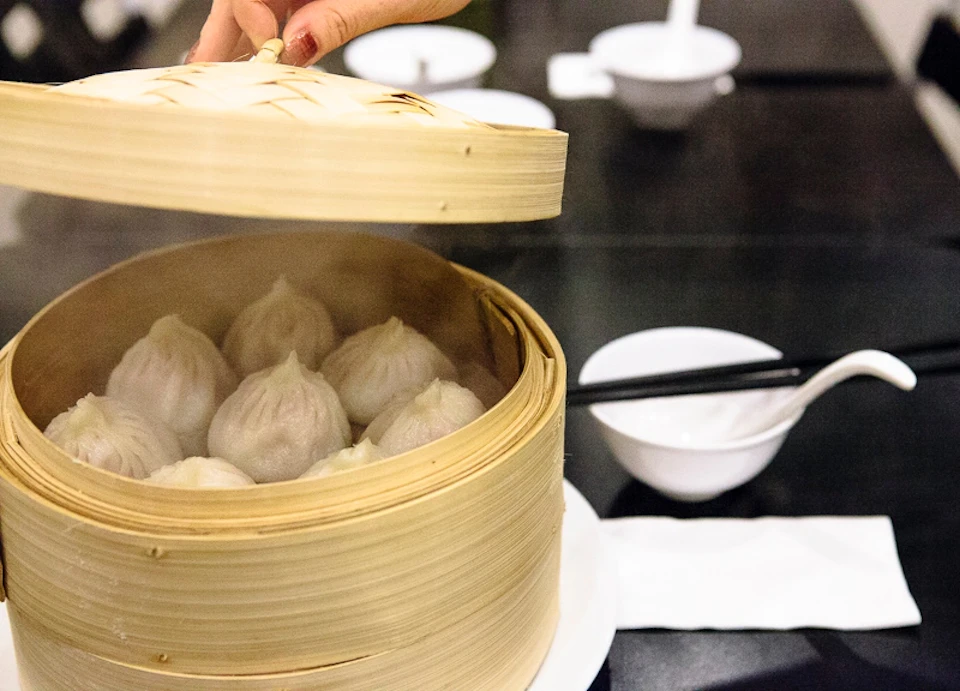 A hand lifting the lid off a bamboo steamer, revealing steamed dumplings. Chopsticks and dishes are on the white tablecloth below.