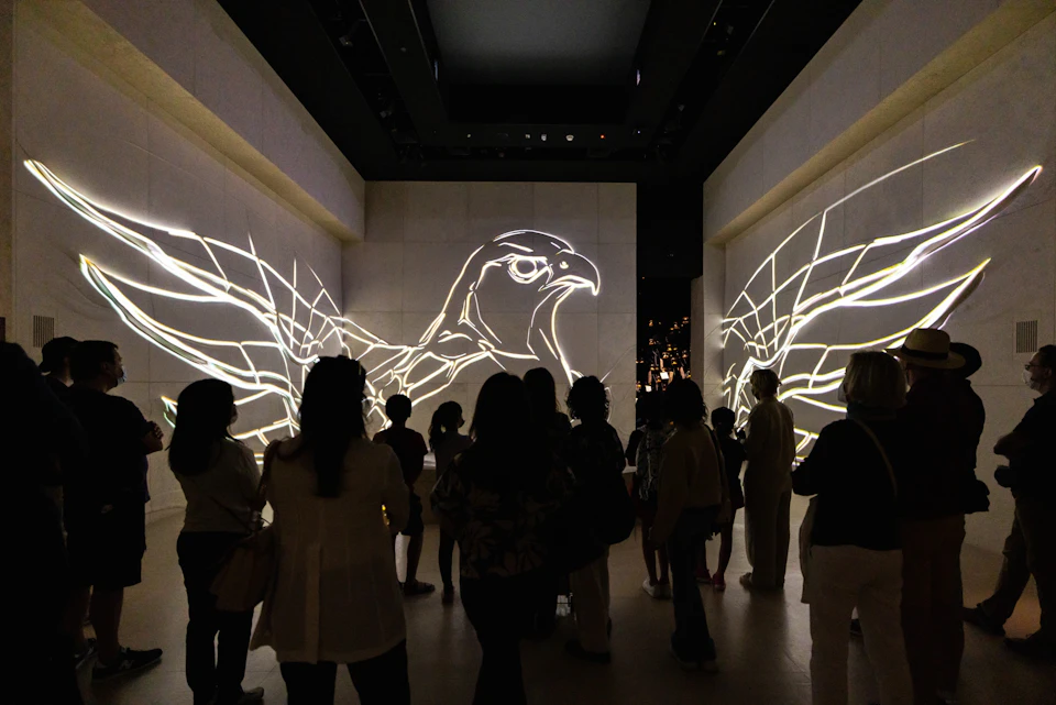 A group of people stand in a dimly lit room with glowing outlines of bird wings and a bird head projected on the walls.