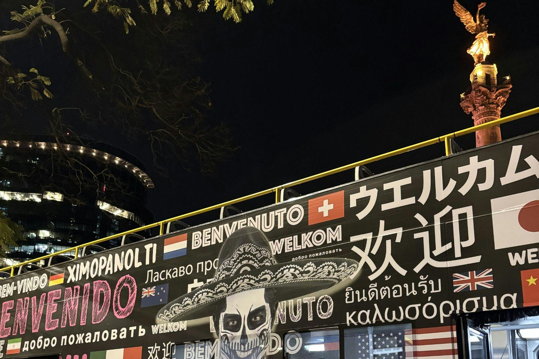 Een levendige muurschildering bij nacht met een schedel in een sombrero en de tekst "Welkom" in meerdere talen. Bomen en gebouwen zijn zichtbaar.