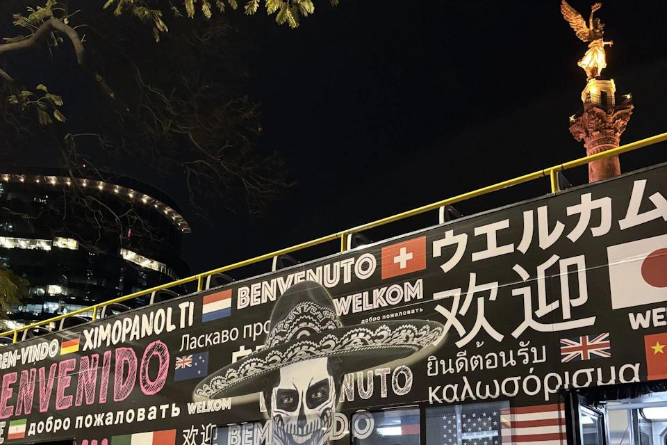 A vibrant mural at night featuring a skull in a sombrero and multiple languages saying "Welcome." Trees and buildings are visible.