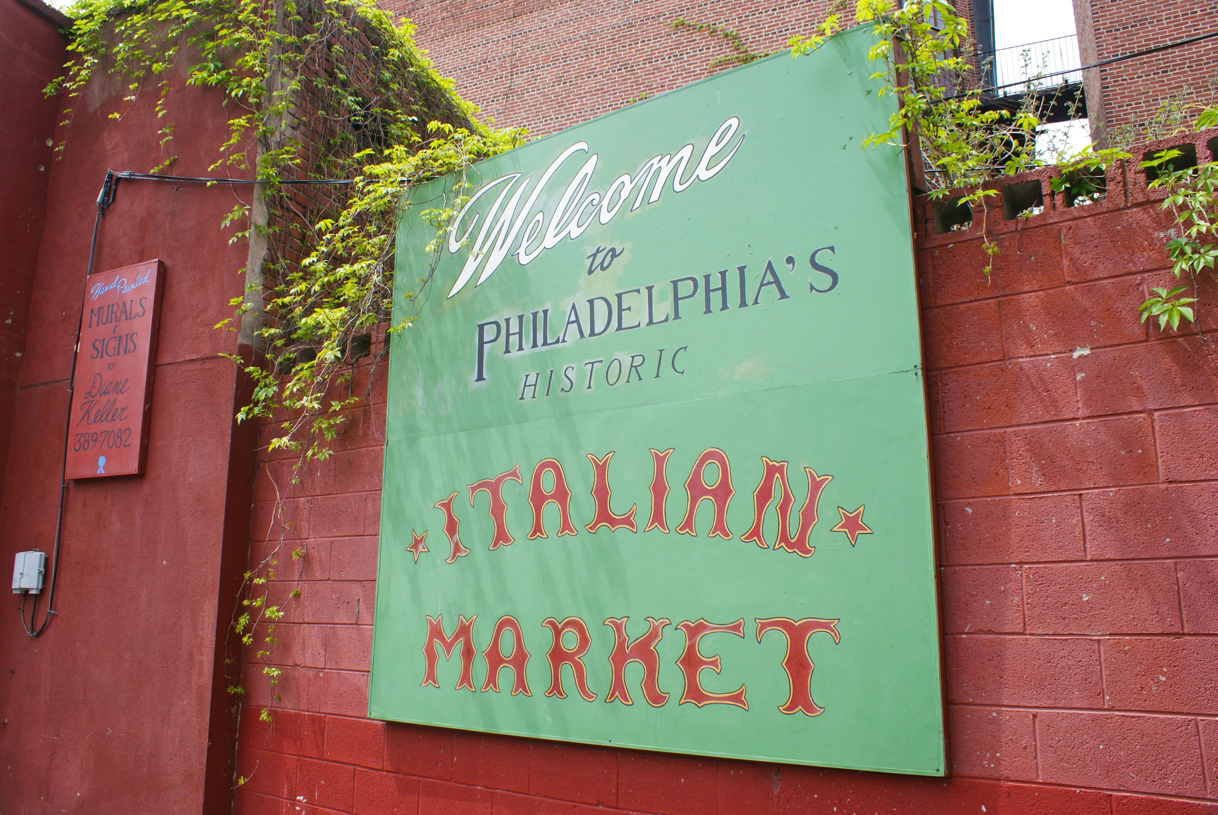 Op een groen bord staat "Welcome to Philadelphia's Historic Italian Market" tegen een rode bakstenen muur met groene wijnranken.