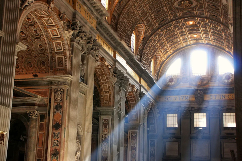 Sunlight streams through ornate windows into a grand interior with elaborately decorated walls and arched ceilings.