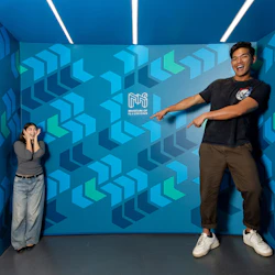 Two people in a room with geometric patterns and the text 'Museum of Illusions' on the wall. One appears much larger.