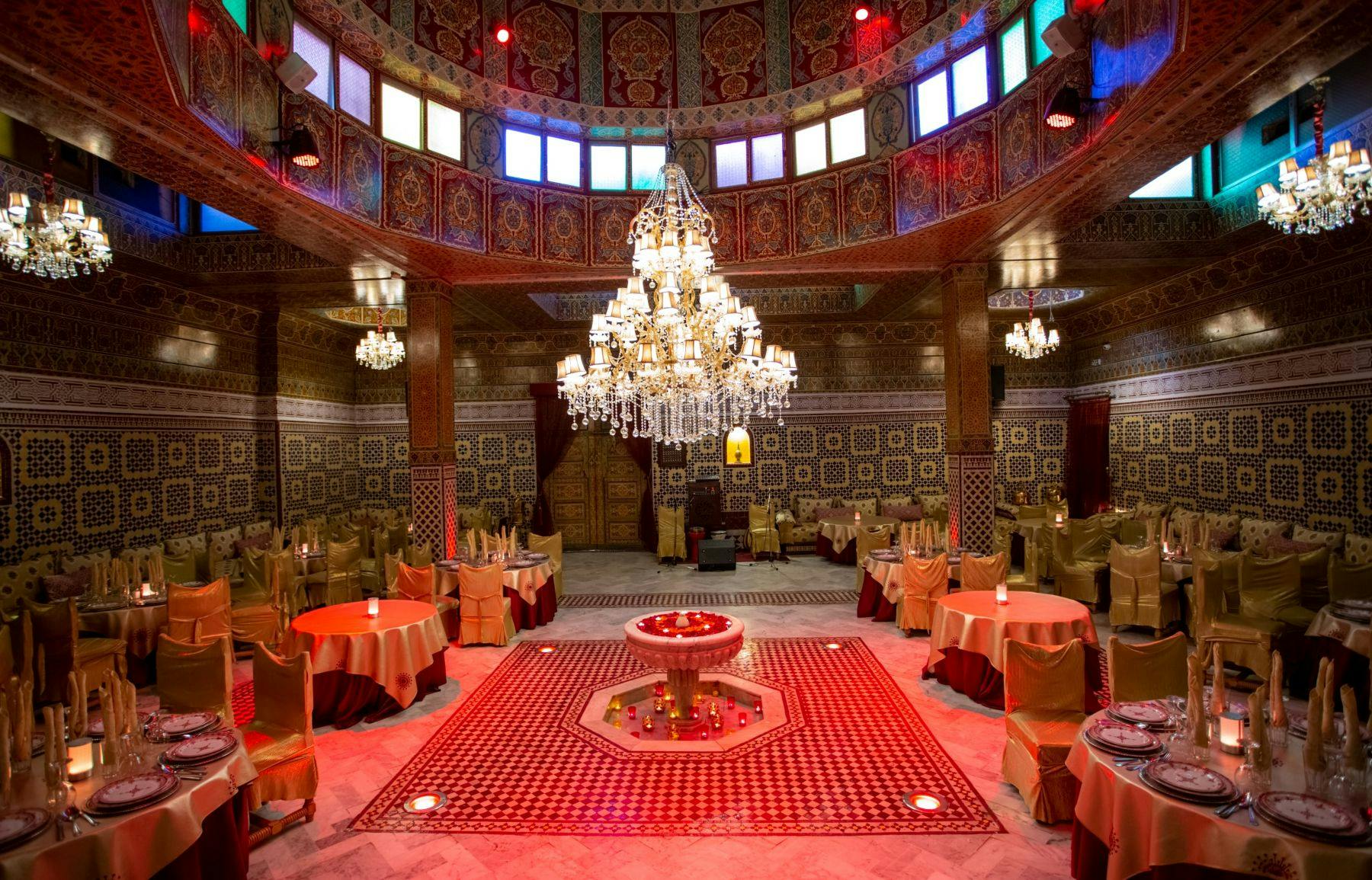 壁と天井が装飾された華やかなダイニングホール、大きなシャンデリア、赤いテーブルクロスの円卓、中央の噴水。