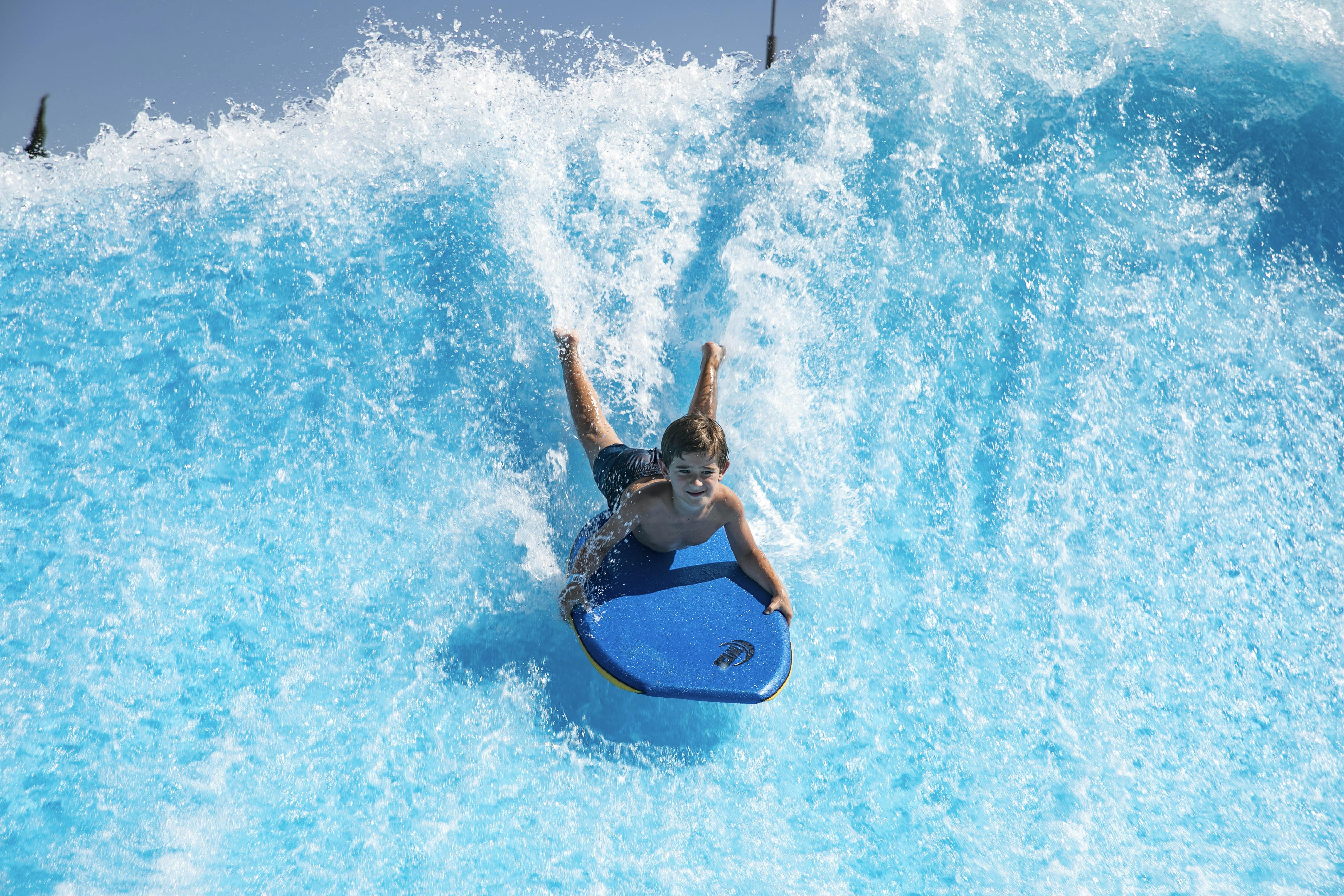 A person rides a bodyboard down a wave in a water park, with blue water splashing around them.