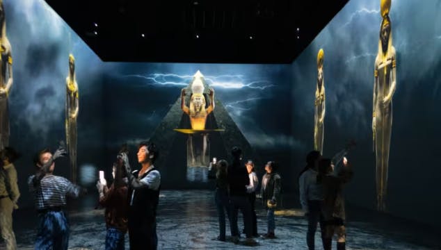 La gente interactúa con una instalación de arte digital que presenta figuras holográficas y una pirámide en una sala oscura.