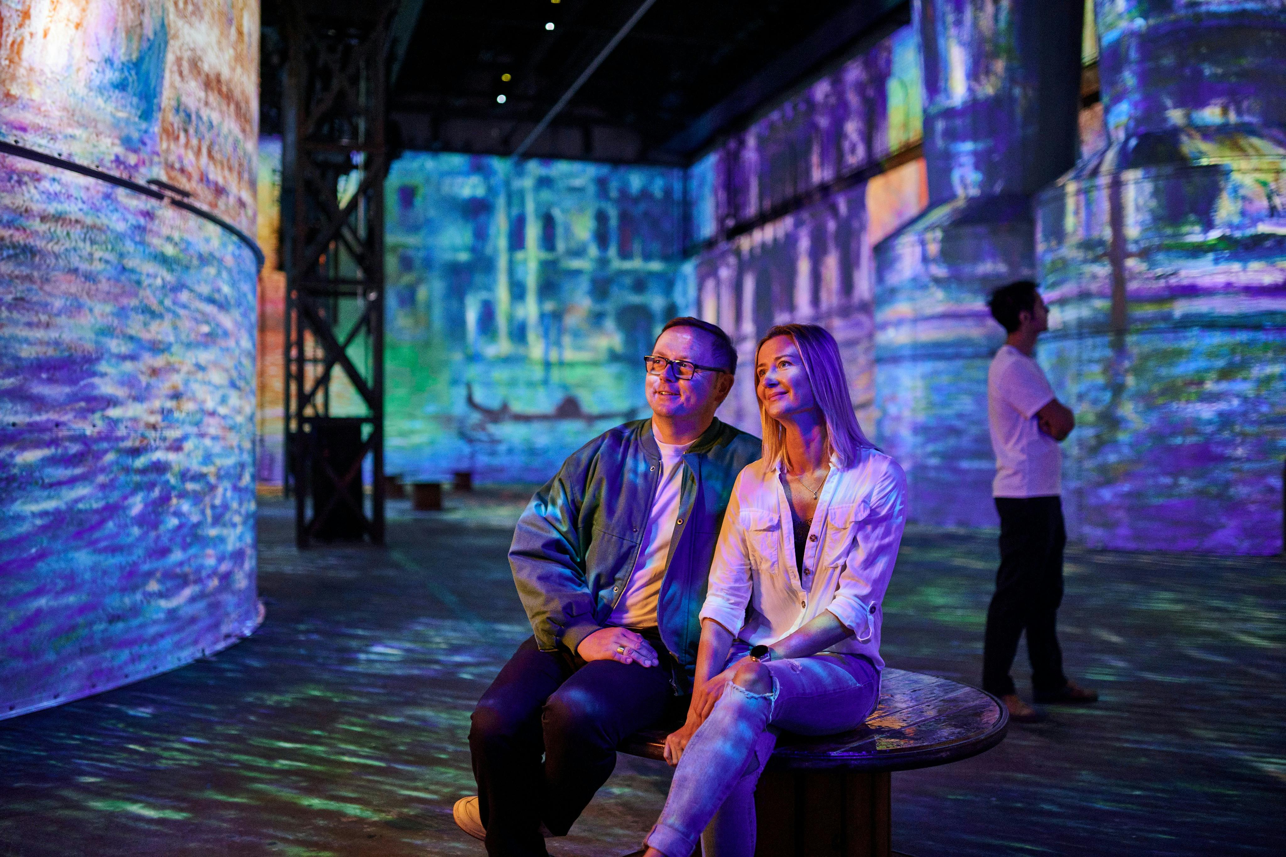 Ένας άνδρας και μια γυναίκα κάθονται σε ένα παγκάκι σε μια εντυπωσιακή έκθεση τέχνης, περιτριγυρισμένοι από πολύχρωμες, προβαλλόμενες εικόνες σε τοίχους και στήλες.