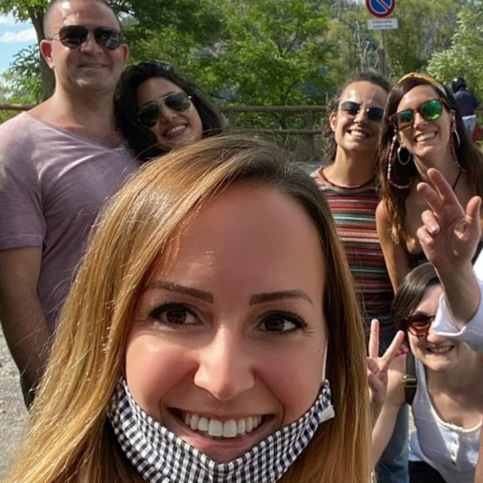 Group of people smiling outdoors, one wearing a face mask and another with a lowered mask, posing for a photo on a sunny day.