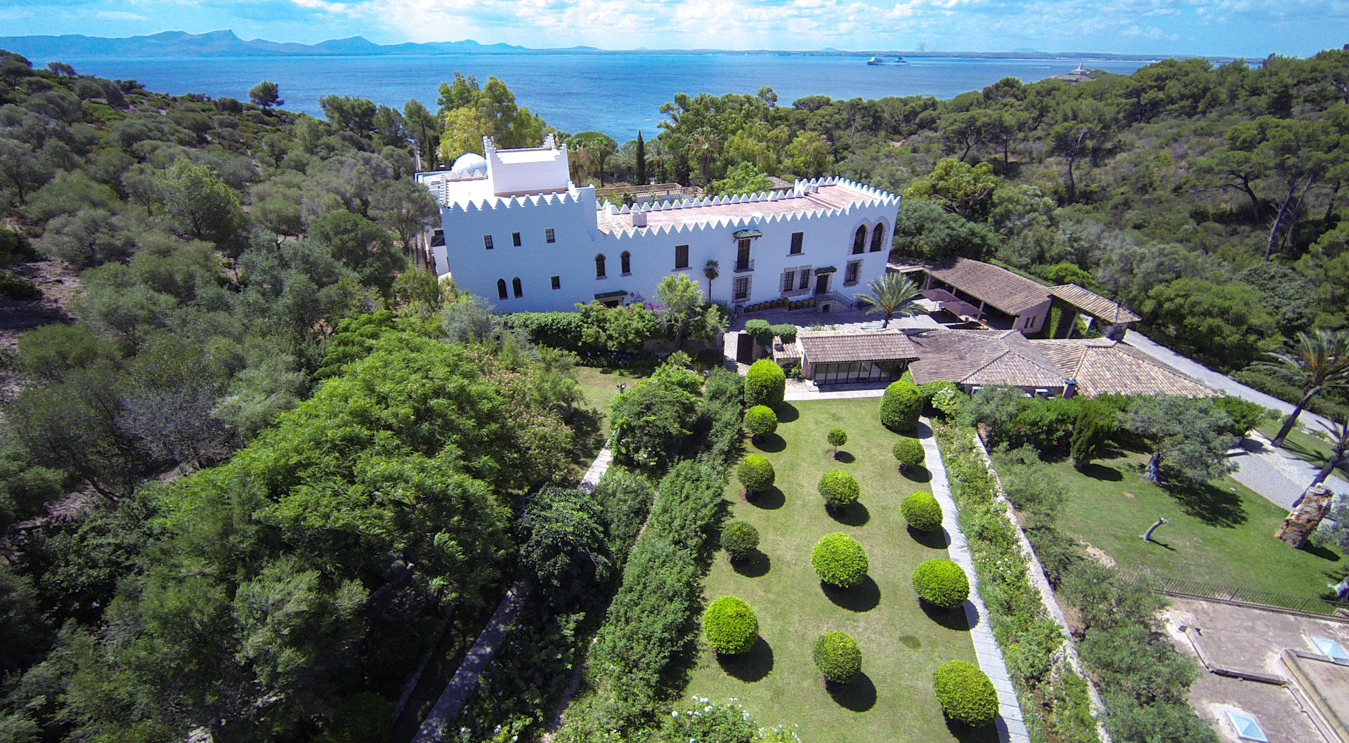Aerial view of a white castle-like building with a manicured garden, surrounded by lush greenery, near the sea.