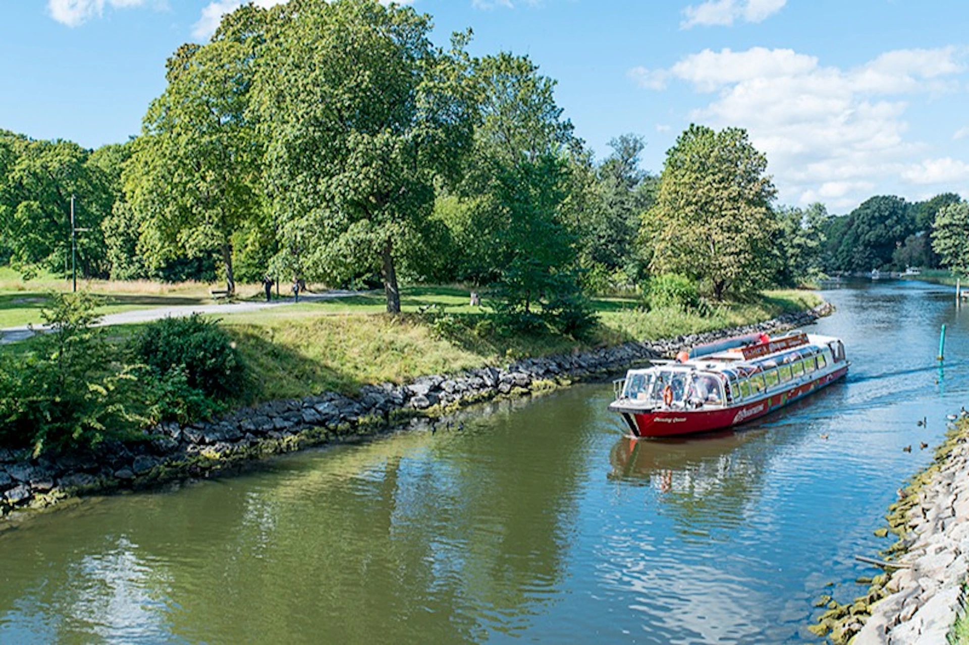 A red tour boat travels down a tree-lined canal on a sunny day with clear skies.