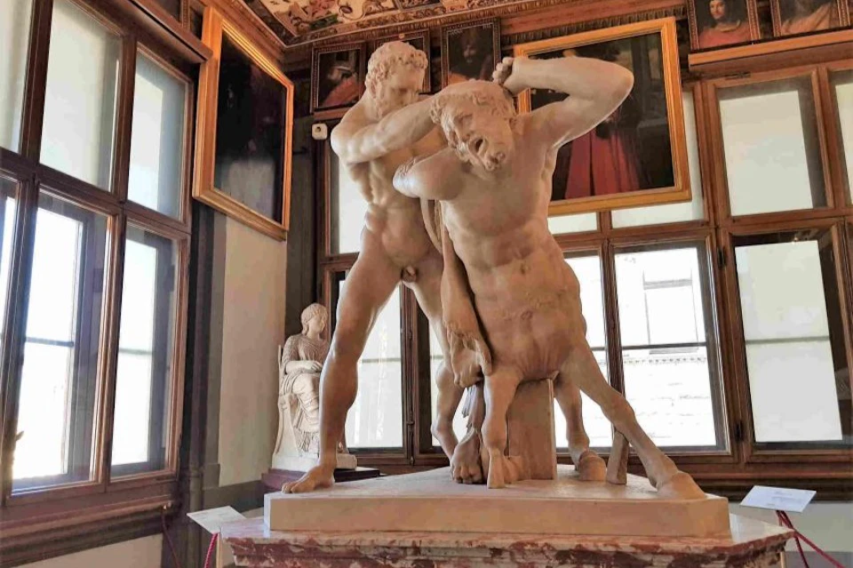 Two marble statues of struggling men in an ornate room with framed paintings and large windows.
