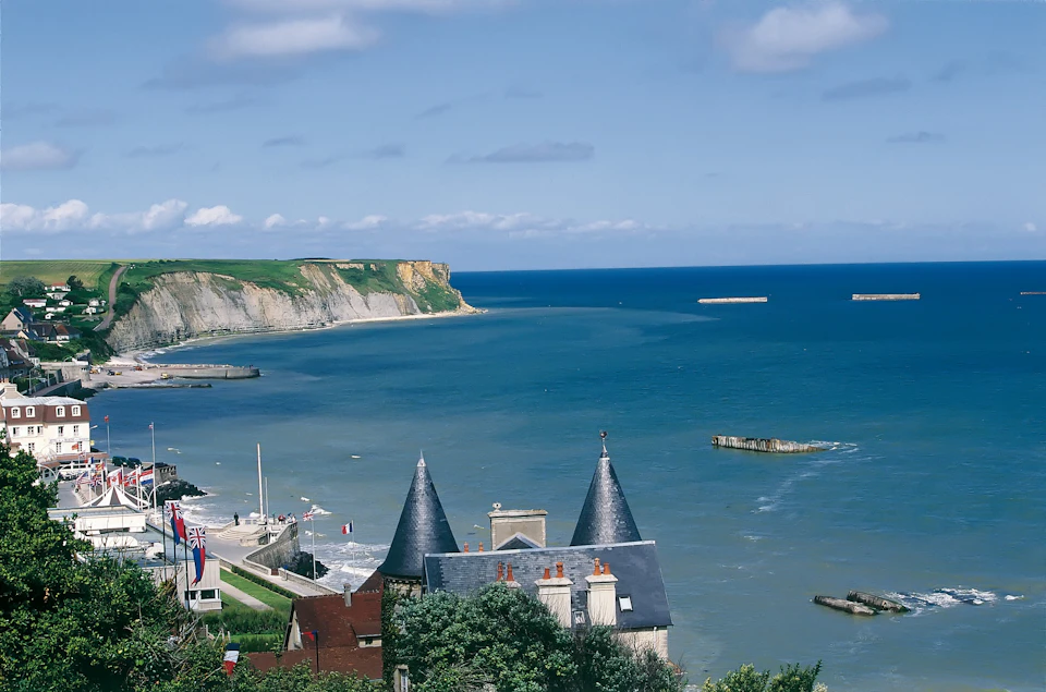 Coastal scene with blue ocean, rocky cliffs in the distance, and a slate-roofed building with pointed towers in the foreground.