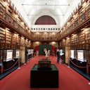 A grand library with high ceilings, filled with shelves of books and display cases. Several people are observing exhibits.