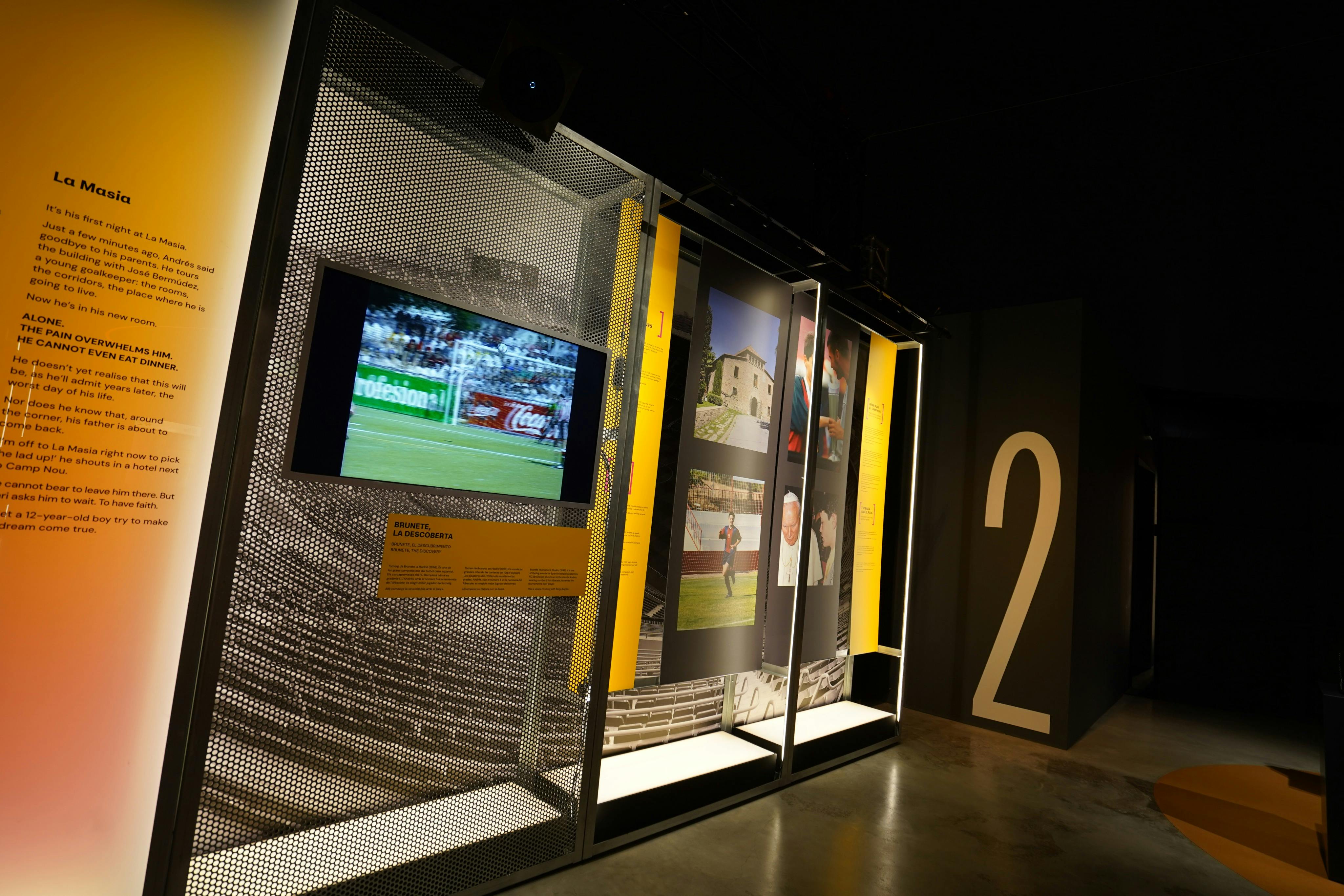 An exhibit in a museum displaying a screen showing a soccer match, photos, and a large number 2 on the wall.