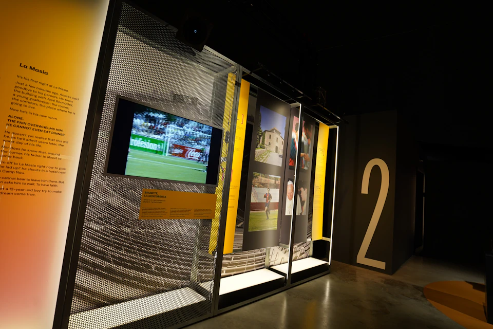 An exhibit in a museum displaying a screen showing a soccer match, photos, and a large number 2 on the wall.
