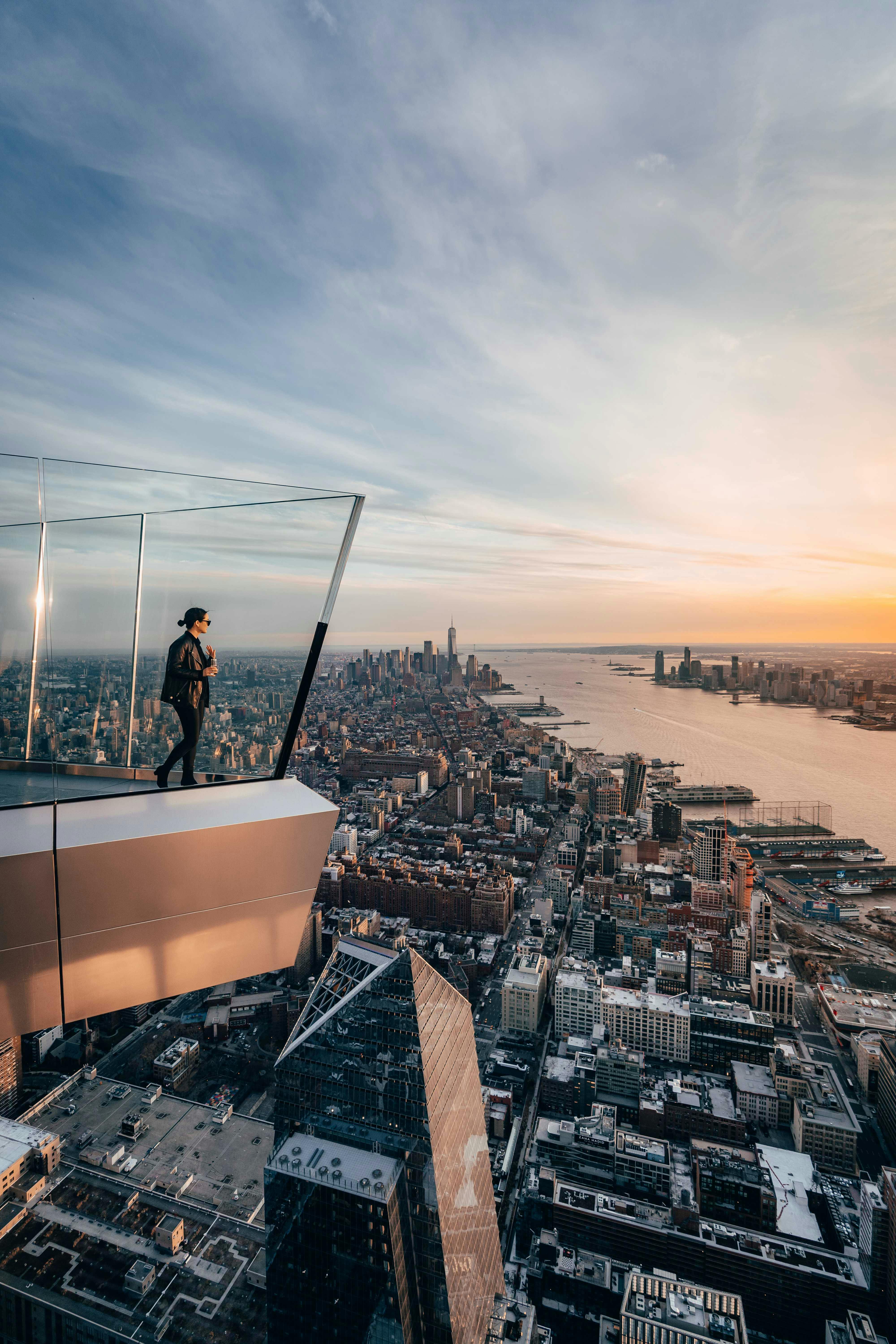 Una persona si trova su una piattaforma di osservazione in vetro sopra una città, con vista su grattacieli, un fiume e un cielo al tramonto.