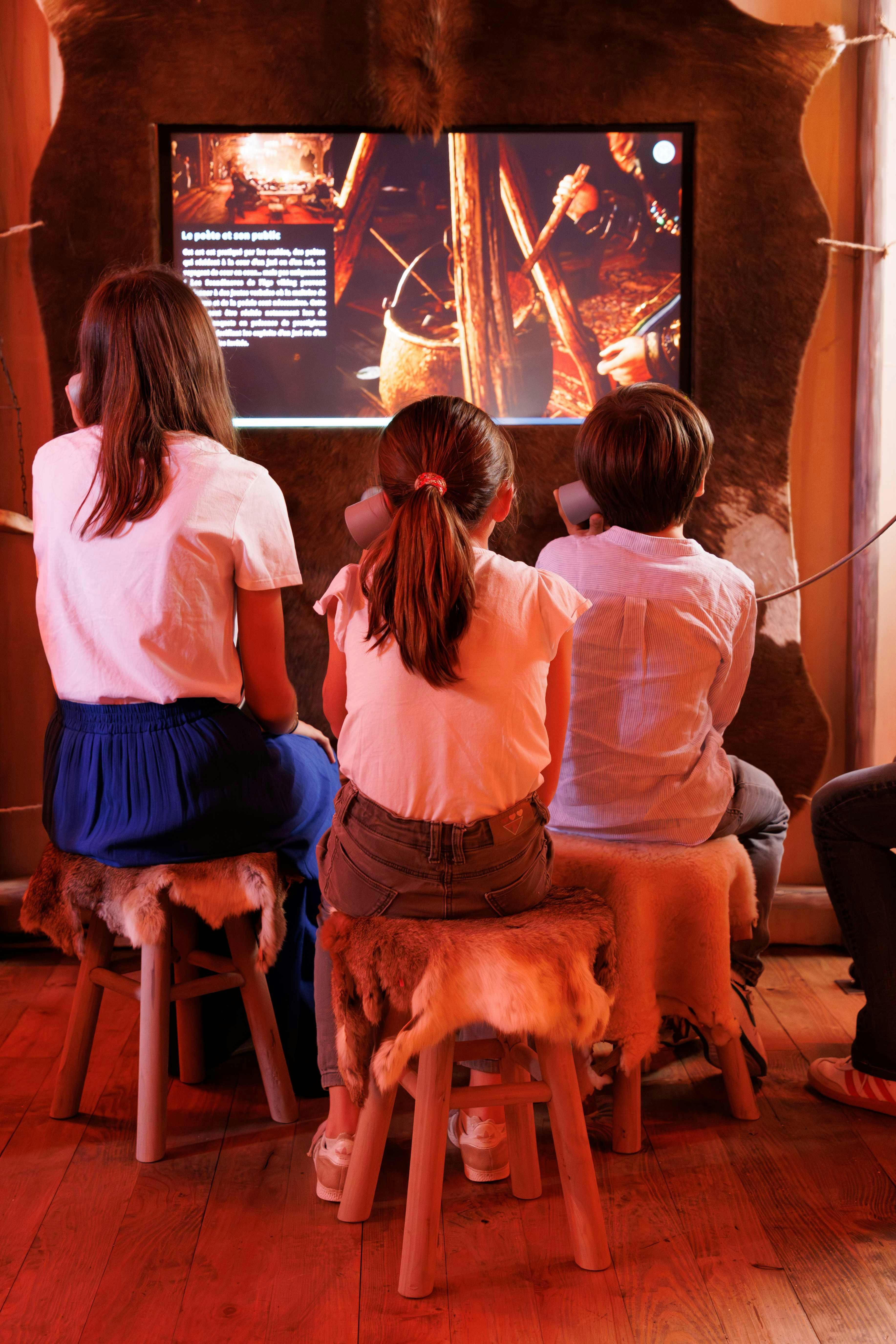 Tre børn sidder på taburetter med dyrehuder, ser en oplyst udstilling og lytter på hovedtelefoner i et svagt oplyst rum.