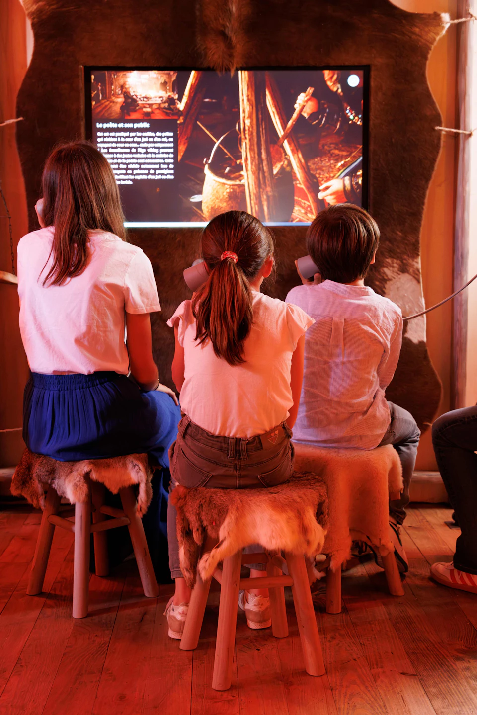 Three children are sitting on stools with animal hide covers, viewing an illuminated exhibit and listening on headphones in a dimly lit room.