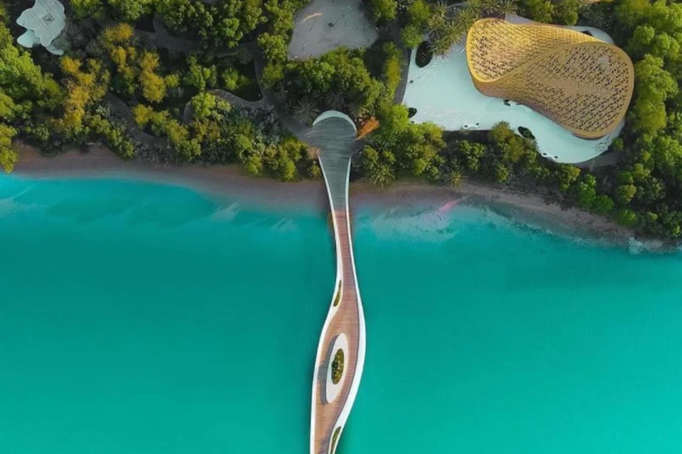 Aerial view of a curving boardwalk extending over turquoise water, bordered by dense greenery and modern architecture.