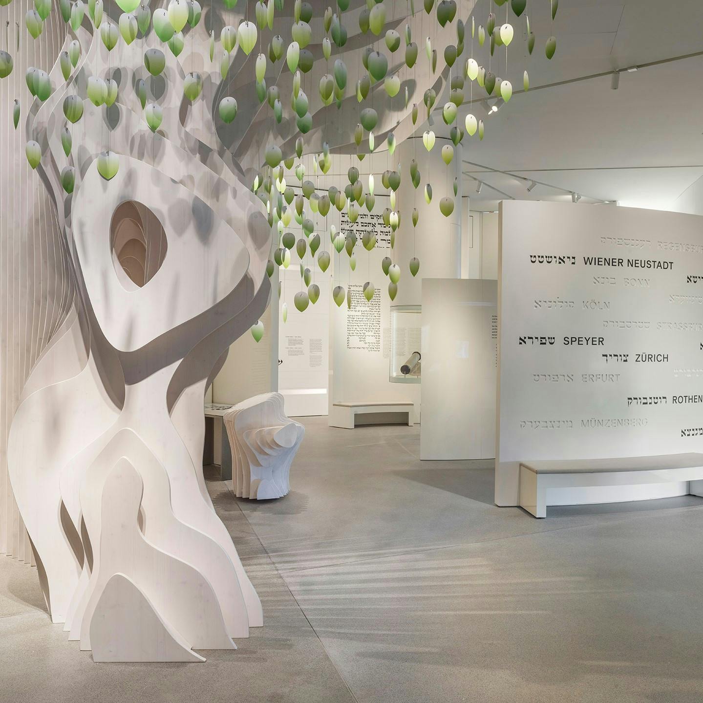 Moderní muzejní expozice se stylizovaným stromem a zavěšenými zelenými listy. Nedaleká zeď zobrazuje názvy měst v hebrejštině a angličtině.