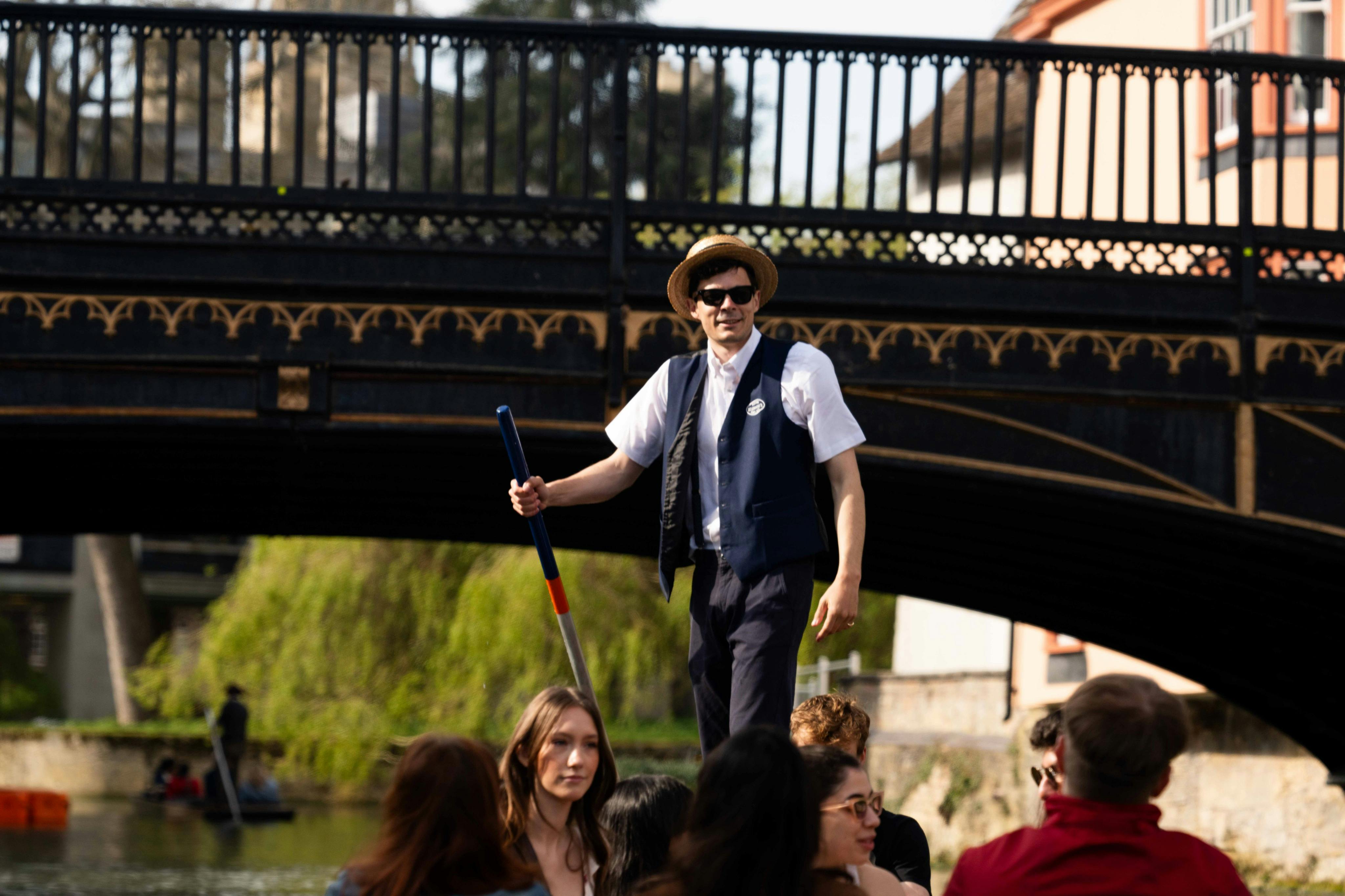Eine Person mit Hut und Sonnenbrille sticht auf einem Fluss unter einer dekorativen Brücke in ein Boot, in dem Menschen sitzen.
