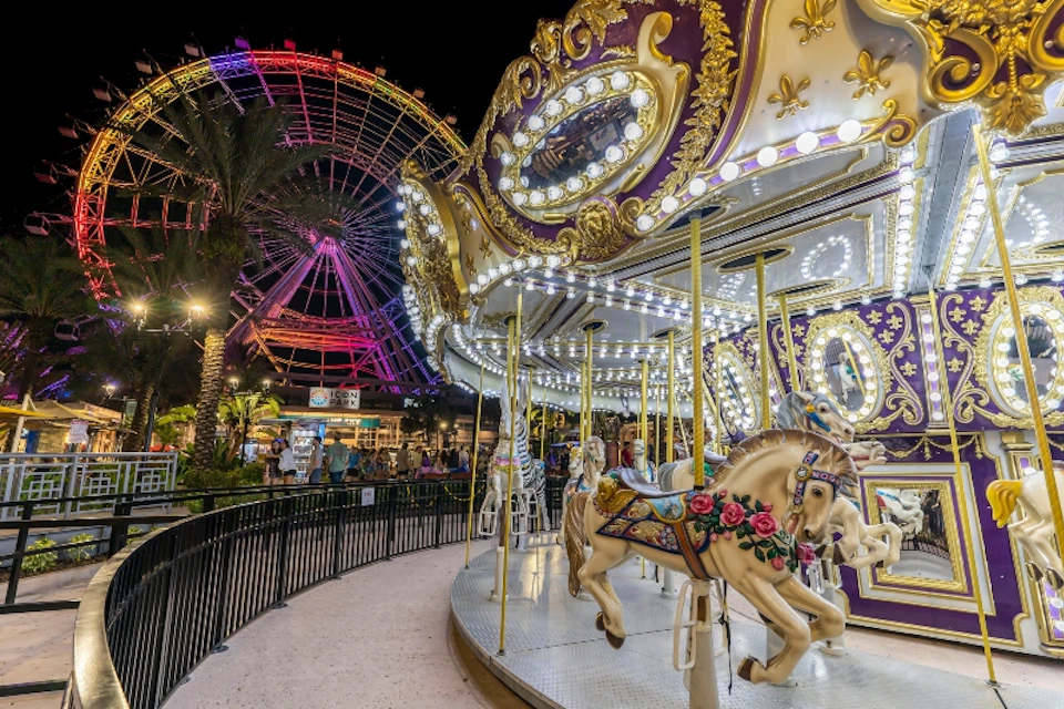 A brightly lit carousel and Ferris wheel at night, with ornate horses in the foreground and a crowd in the background.