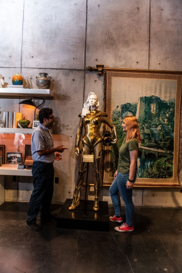 装飾の棚があり、壁にタペストリーが飾られた部屋の中で、黄金の戦士像の横に2人の人物が立っています。