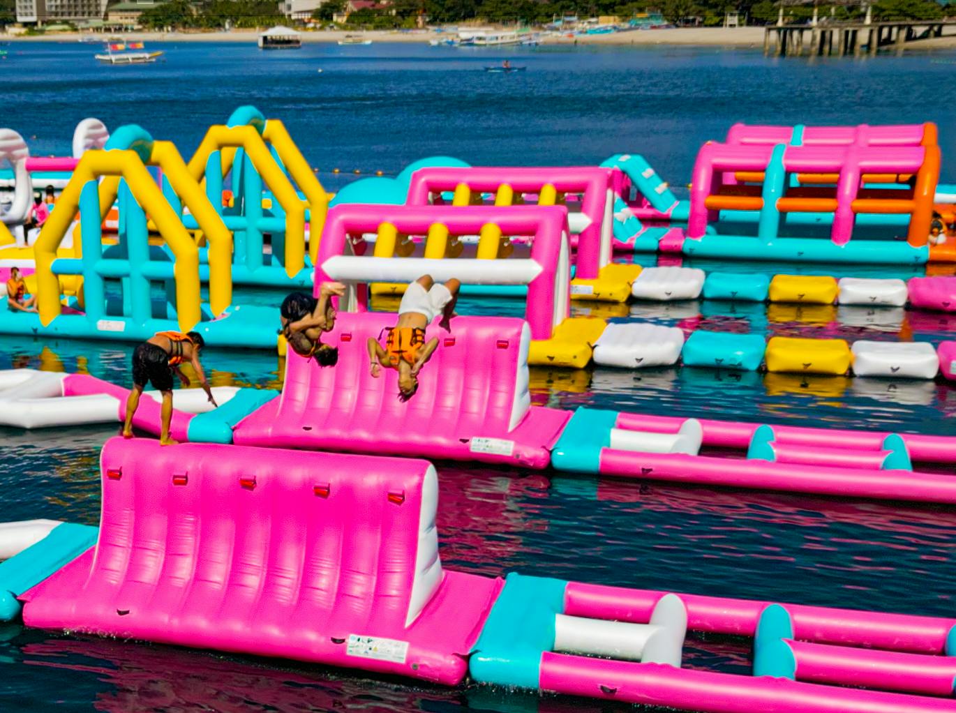 People playing on a colorful inflatable water park with slides and obstacles, floating on a lake.