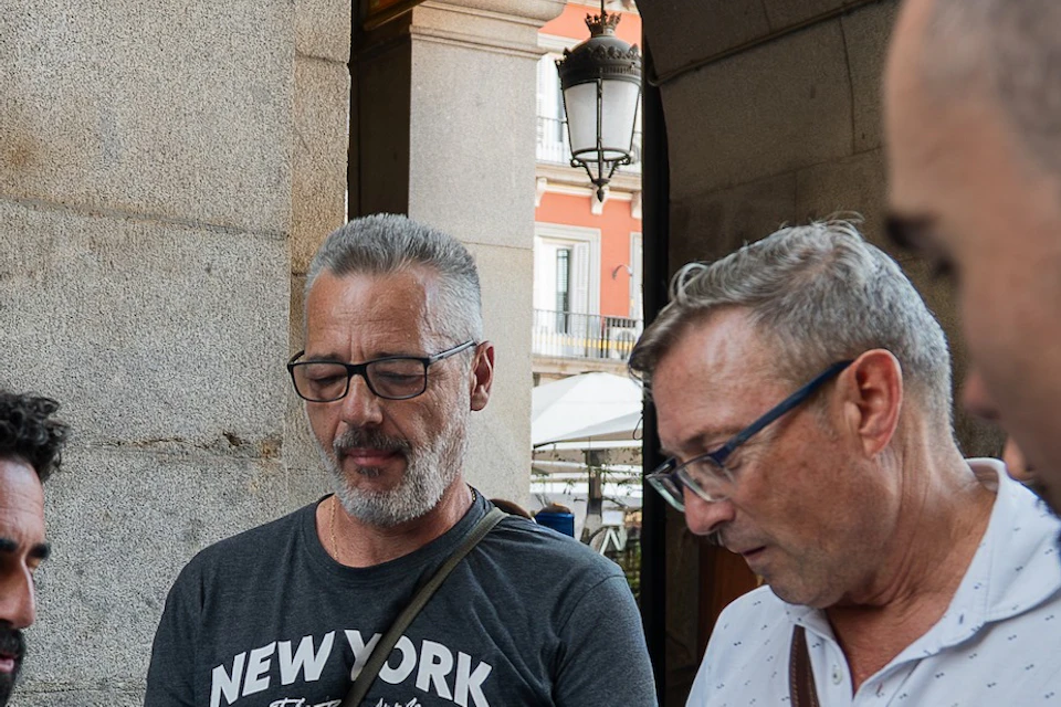 Four men are standing together outside, looking at a document or map, with a building and a lamp post visible in the background.