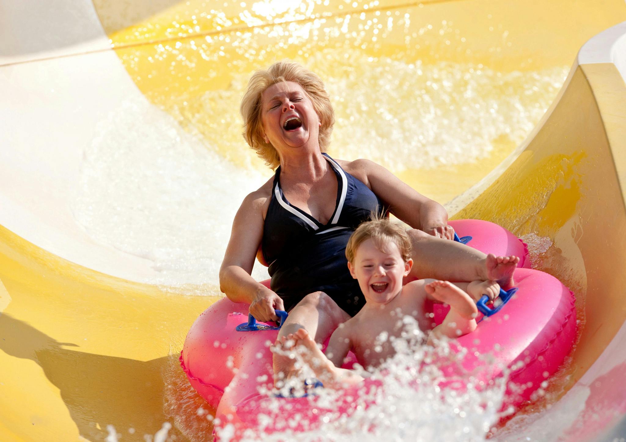 Een vrouw en een kind in een roze opblaasbare ring glijden lachend van een waterglijbaan.