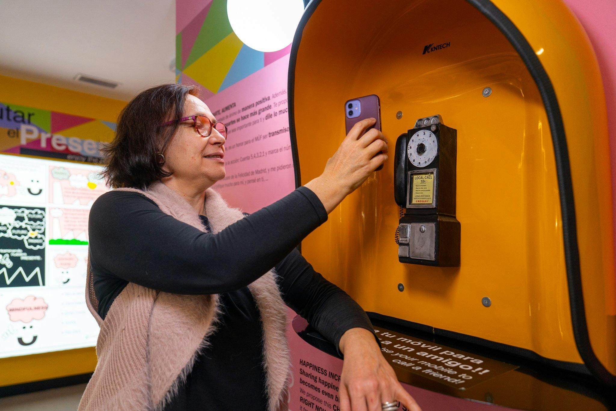 Μια γυναίκα βγάζει μια φωτογραφία με το τηλέφωνό της σε μια εσωτερική έκθεση με πολύχρωμους τοίχους και ενημερωτικές οθόνες.