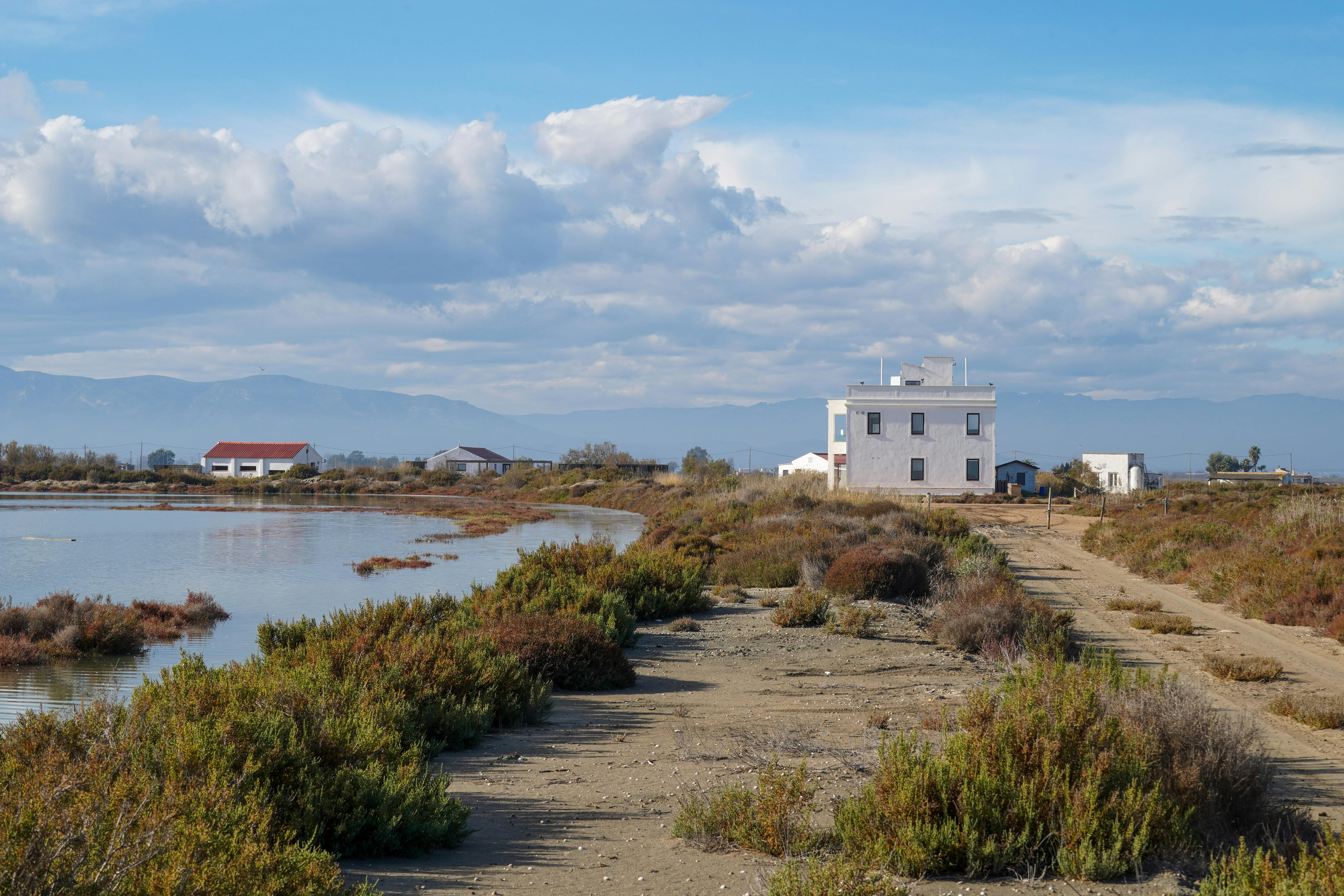 Krajina u jezera s budovami, prašnými cestami, keři a horami pod částečně zataženou oblohou.
