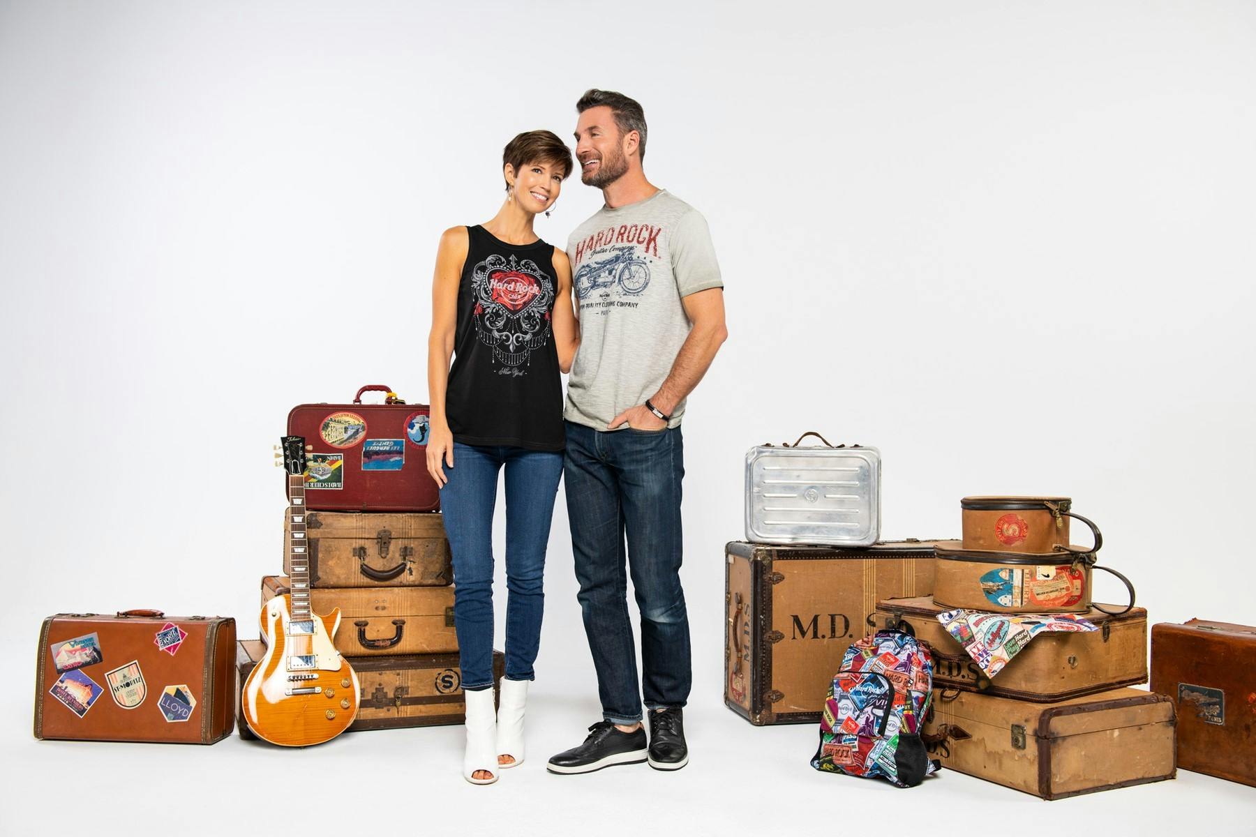 A smiling couple stands next to vintage suitcases, a guitar, and a colorful backpack against a white background.