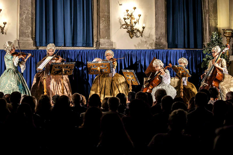 Four musicians in period costumes perform with string instruments on a stage in front of an audience. Blue curtains and dim lighting.