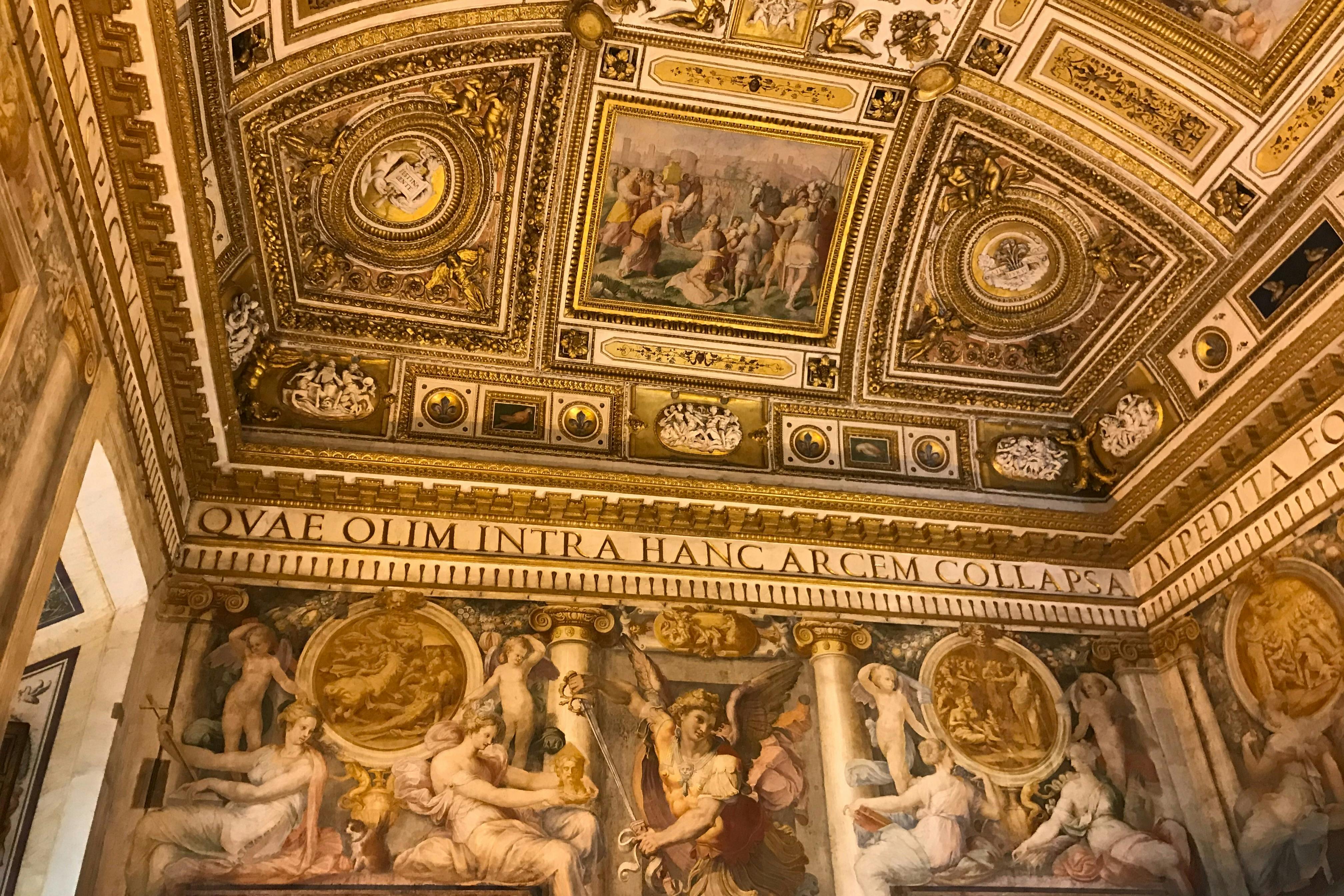 Verschnörkelte Decken- und Wandfresken mit detaillierten Kunstwerken und lateinischen Inschriften, die klassische und religiöse Themen darstellen.