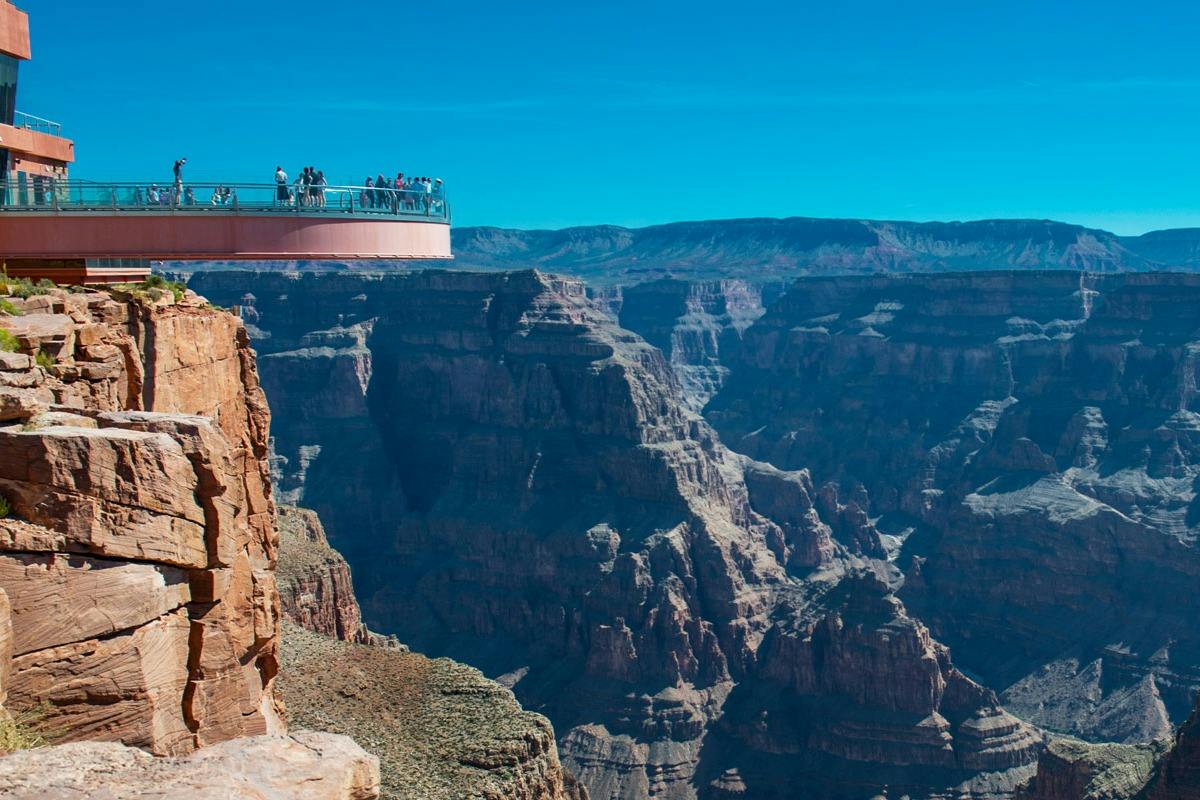 A glass observation deck extends over a deep canyon with visitors viewing the expansive rocky landscape under a clear blue sky.
