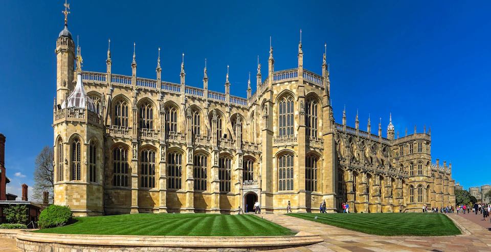 A gothic-style stone building with large windows and tall spires, surrounded by well-manicured grass and a few people walking.