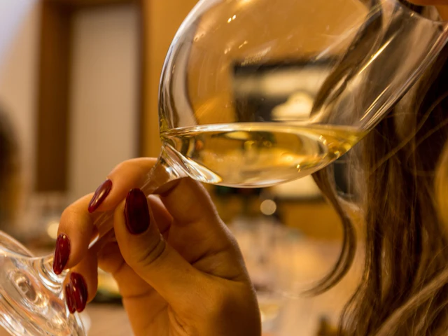 A person holding a glass of white wine close to their face, indoors with a blurred background.