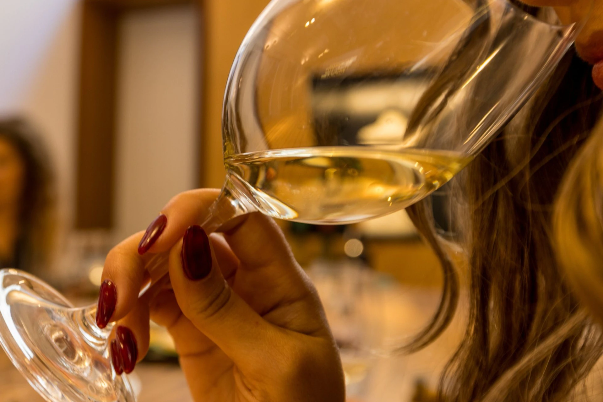 A person holding a glass of white wine close to their face, indoors with a blurred background.