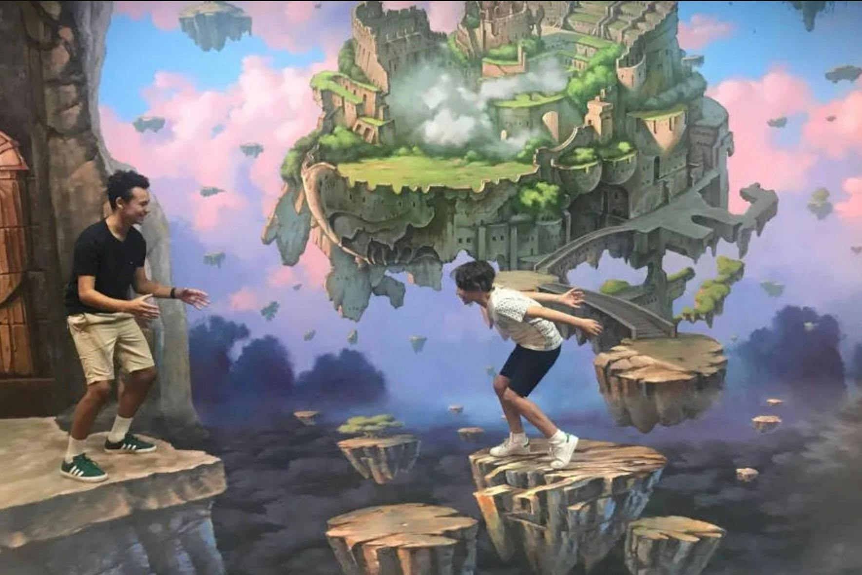 Two people interact in front of a painted scene of floating islands and cliffs, creating an optical illusion.