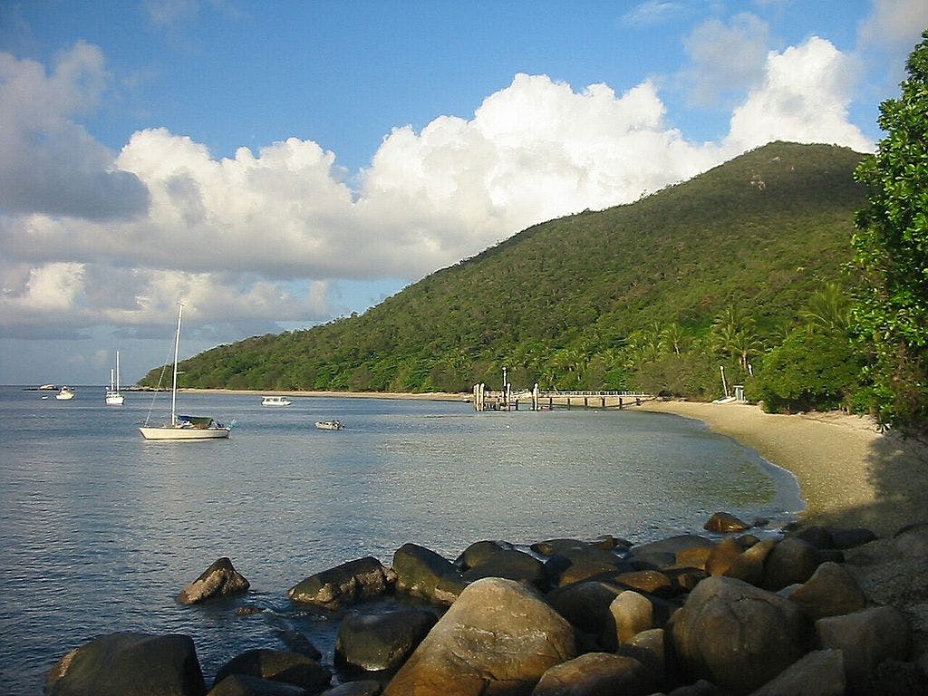 Una serena scena di spiaggia con primo piano roccioso, acque calme con barche ancorate, un molo e una collina coperta di alberi sotto un cielo parzialmente nuvoloso.