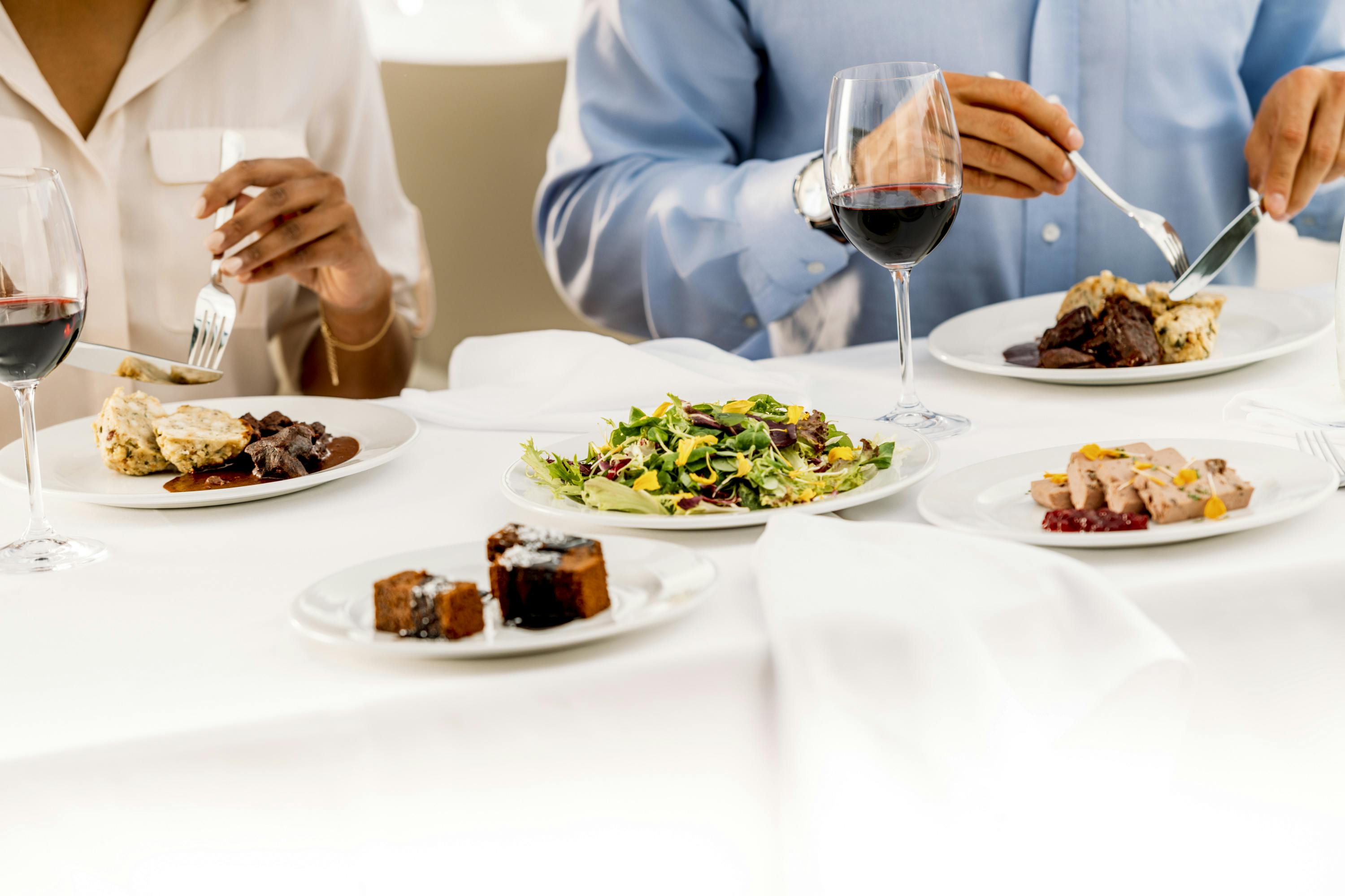 Two people dining with plates of salad, meat dishes, desserts, and a glass of red wine on a white tablecloth.