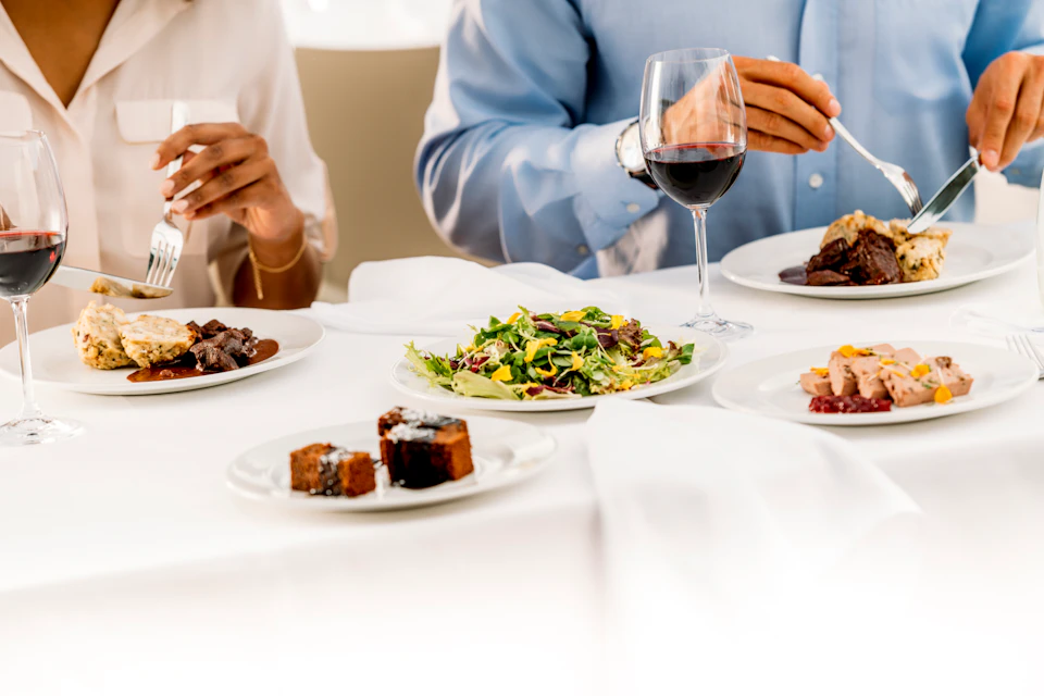 Two people dining, eating salad, meat, and dessert, with a glass of red wine on a white tablecloth.