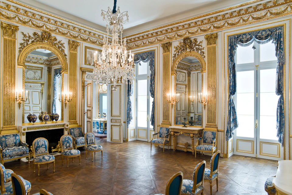 Ornate room with gold trim, a large chandelier, blue and gold upholstered chairs, and mirrored wall panels with intricate detailing.