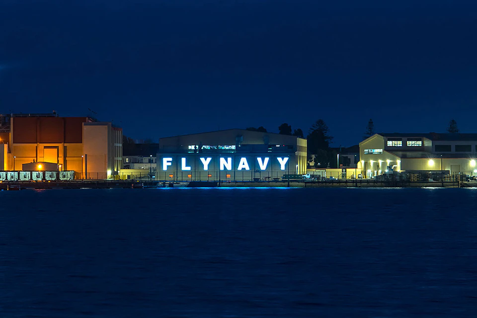 A building by the water with "FLYNAVY" in large, illuminated letters at night. Nearby buildings and trees are dimly lit.