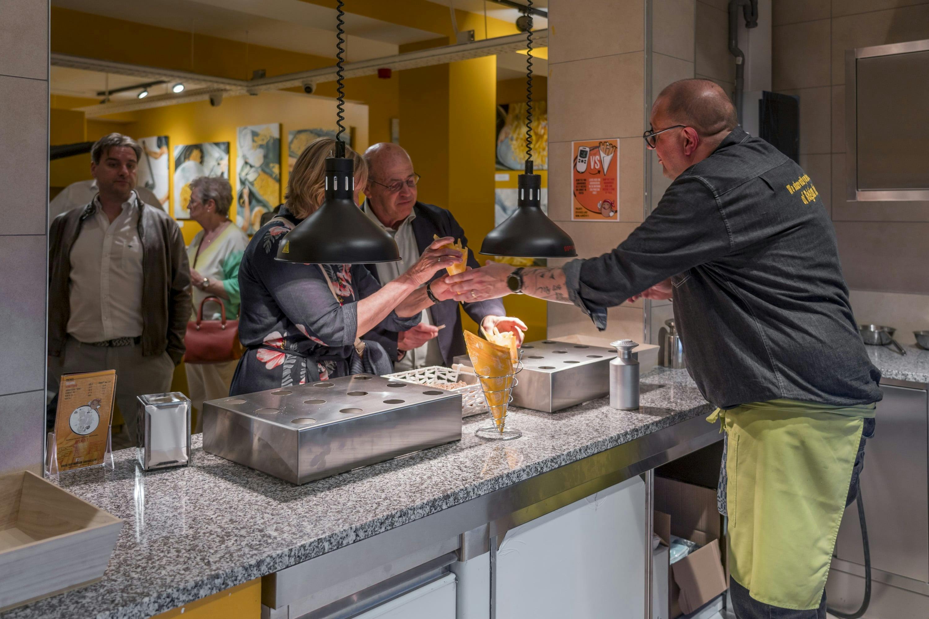 Ludzie czekają przy ladzie; mężczyzna podaje rożki z jedzeniem kobiecie pod wiszącymi światłami w jasno oświetlonym pokoju.