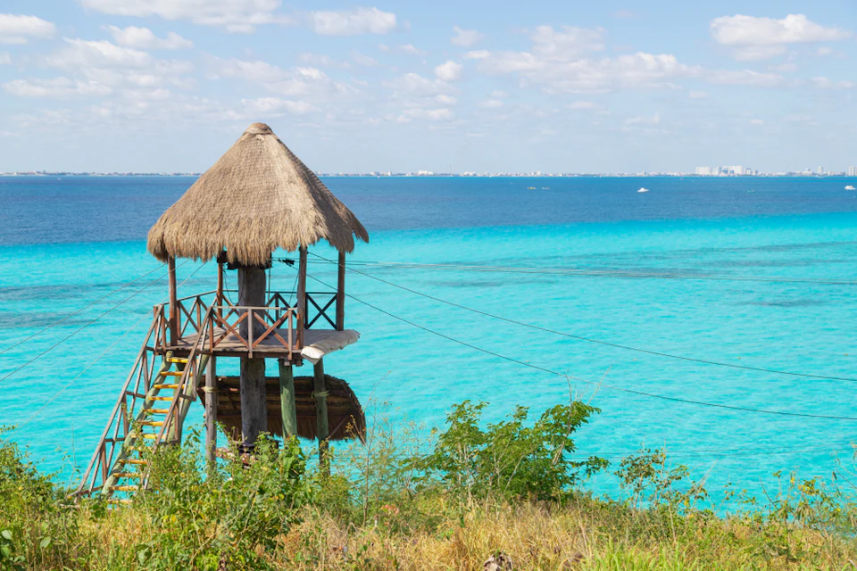 A wooden hut with a thatched roof stands near turquoise ocean waters, surrounded by greenery and a distant city skyline.