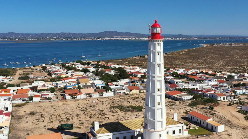 A tall white lighthouse with a red top stands near a cluster of houses by the water with hills in the background.