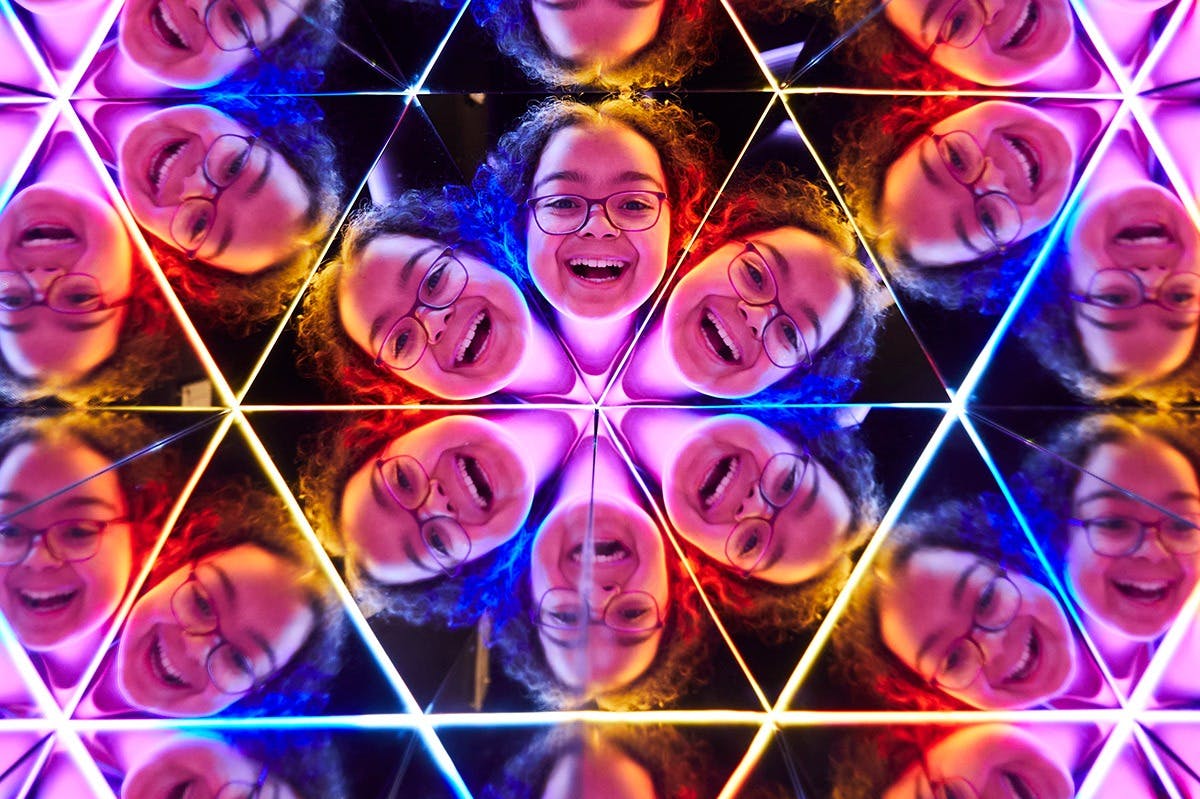 Un visage souriant avec des lunettes se reflète plusieurs fois dans un kaléidoscope à miroirs avec des effets de lumière colorés.